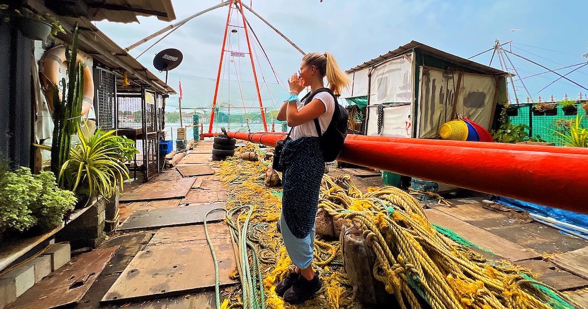 Blonde lady dressed in loose blue trousers, trainers, a white t-shirt and kimono on a Chinese fishing net in Cochin