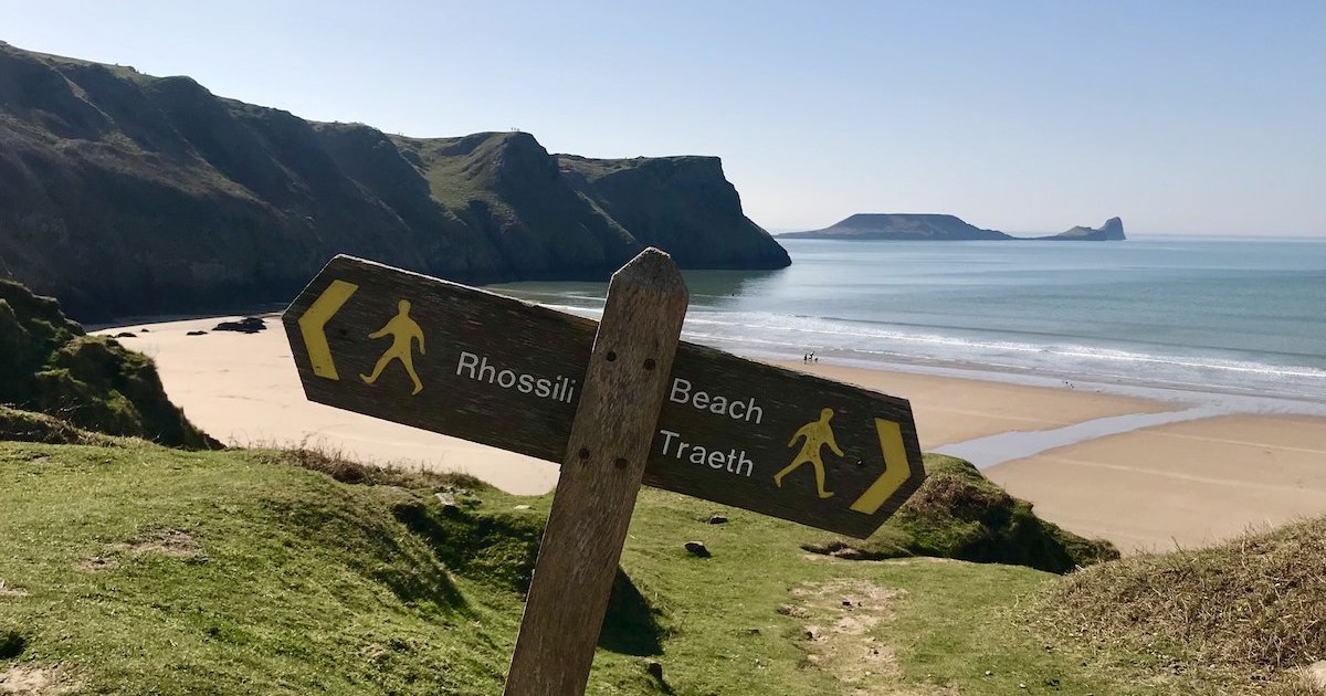A wooden signpost for Rhossili village and Rhossili Beach with Worm's Head in the background.