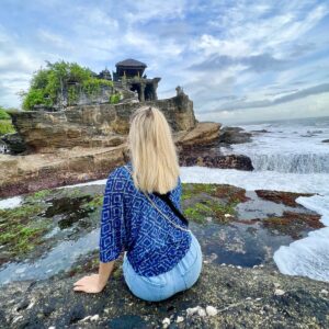 Blonde woman wearing a blue shawl sits on the rocks in front of Tanah Lot Temple at sunset.