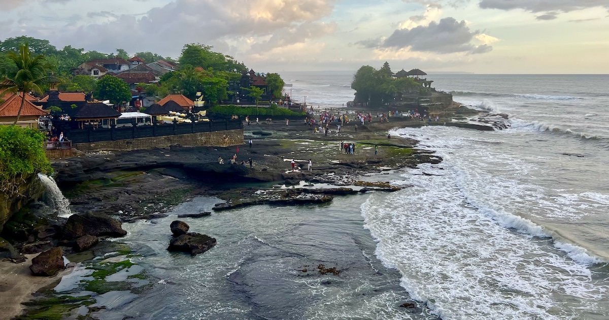 A curved beach with a waterfall curves towards Tanah Lot Temple at sunset.