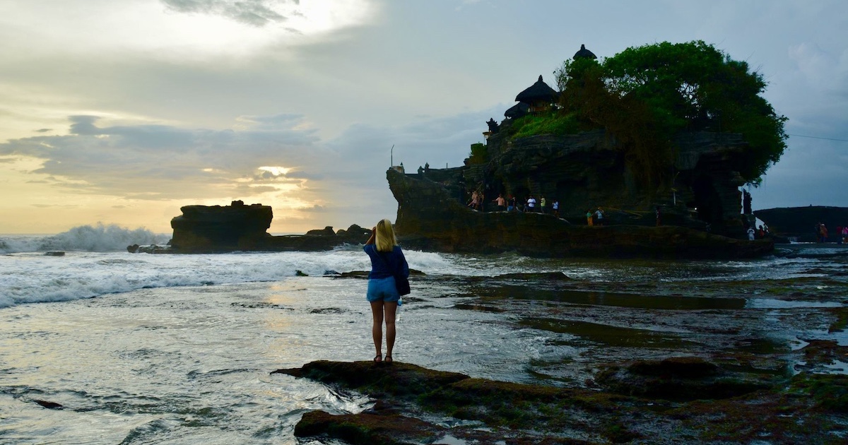 Blonde woman stands on a rock on Sunset Beach at a Tanah Lot sunset.