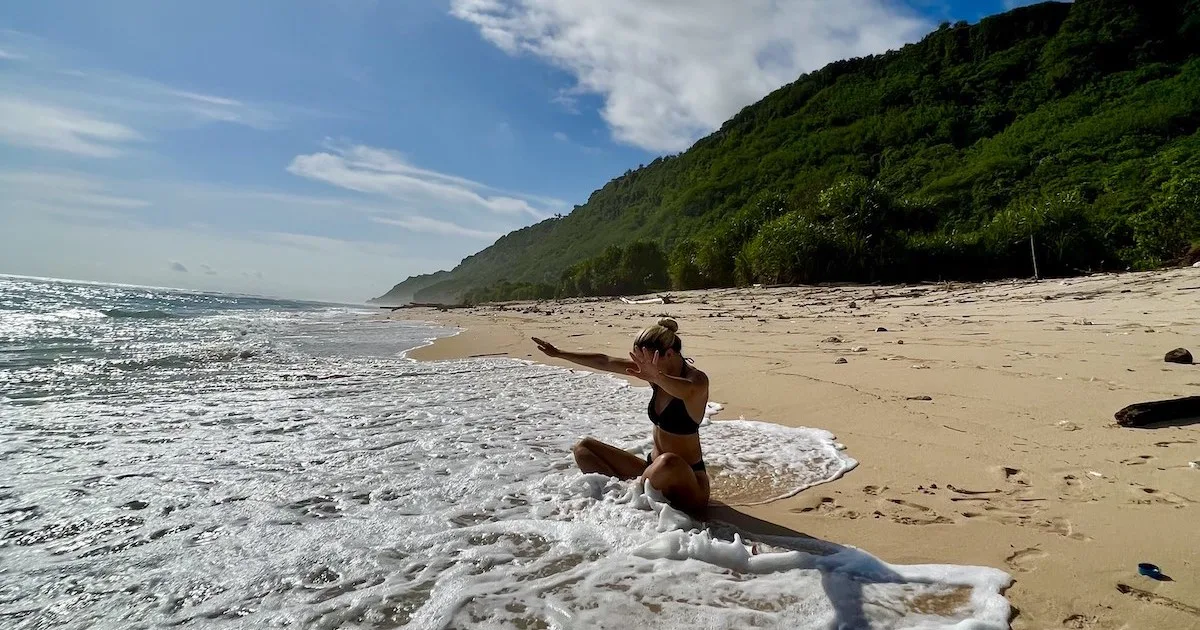 Girl sits cross-legged in the small waves on Nunggalan Beach, raising her arms.