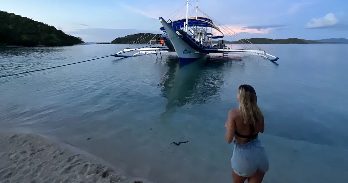 Girl wearing denim shorts and a bikini shorts stands on the beach next to a bangka boat in Linapacan