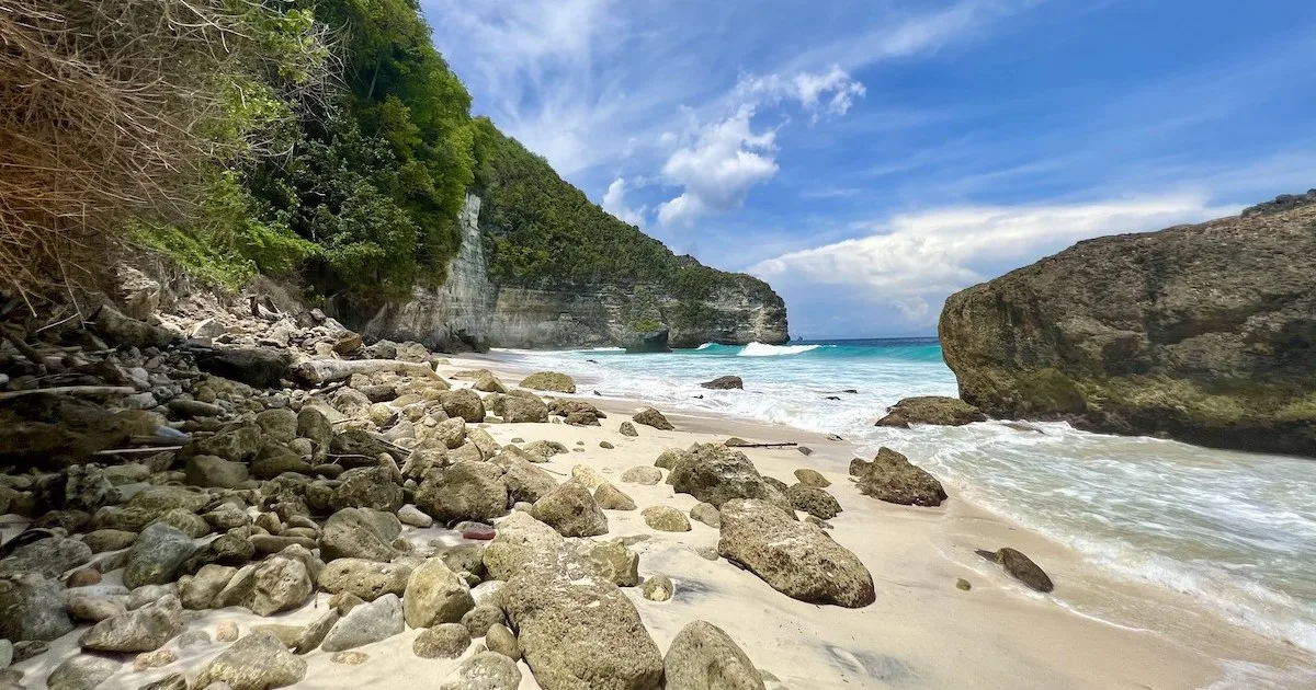 Sparse rocks lead to an empty sandy beach with limestone cliffs.