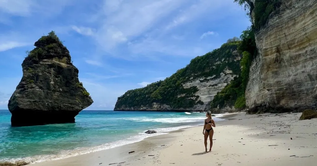 Woman wearing a bikini stands on sandy Suwehan Beach, looking at a diamond-shaped rock jutting out of the sea.