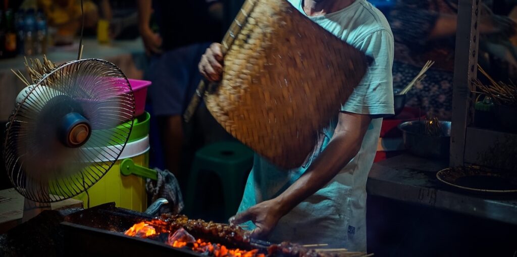 Balinese local waves a bamboo mat over a barbecue cooking chicken skewers/