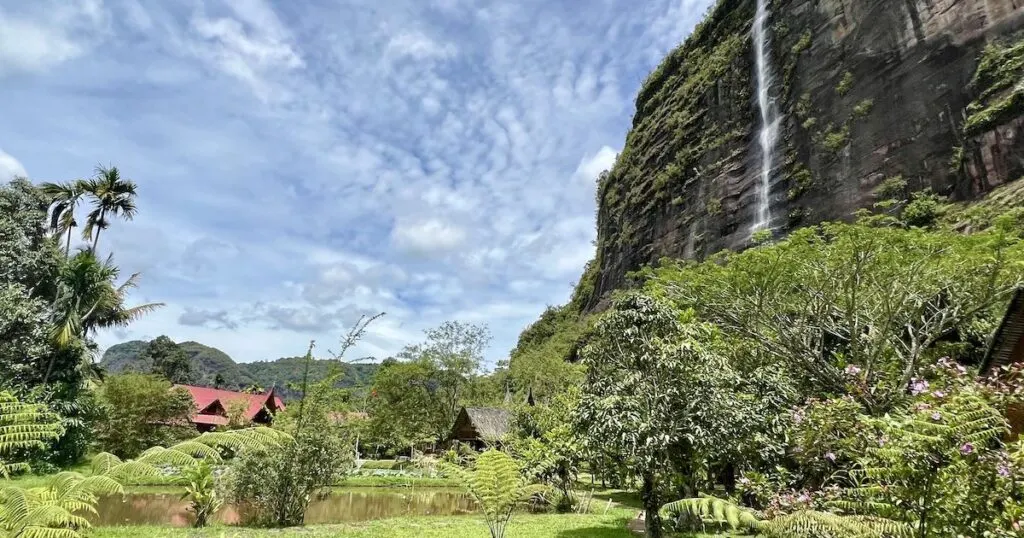 Abdi Homestay's wooden chalets framed by a cliff and waterfall in Harau Valley.