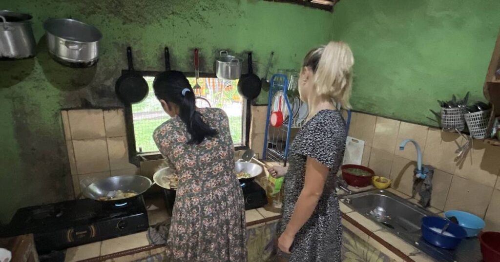 Tourist wearing a t-shirt dress and cooking Indonesian cuisine in a basic kitchen at Abdi Homestay in Harau Valley.