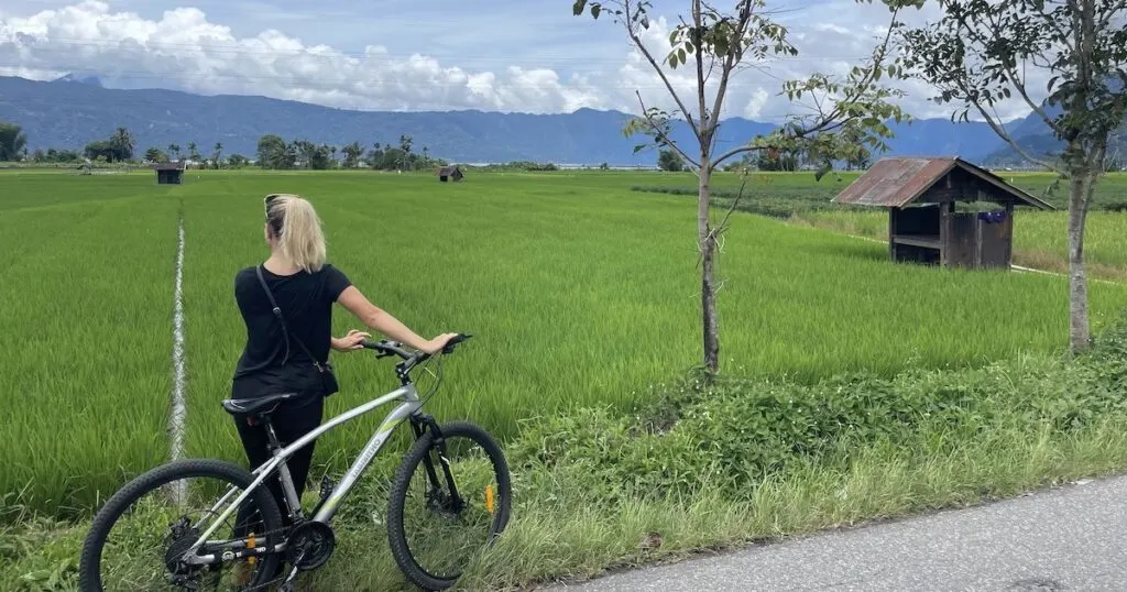 Girl wearing a black t-shirt and black linen trousers holds a bicycle next to rice fields at Lake Maninjau.