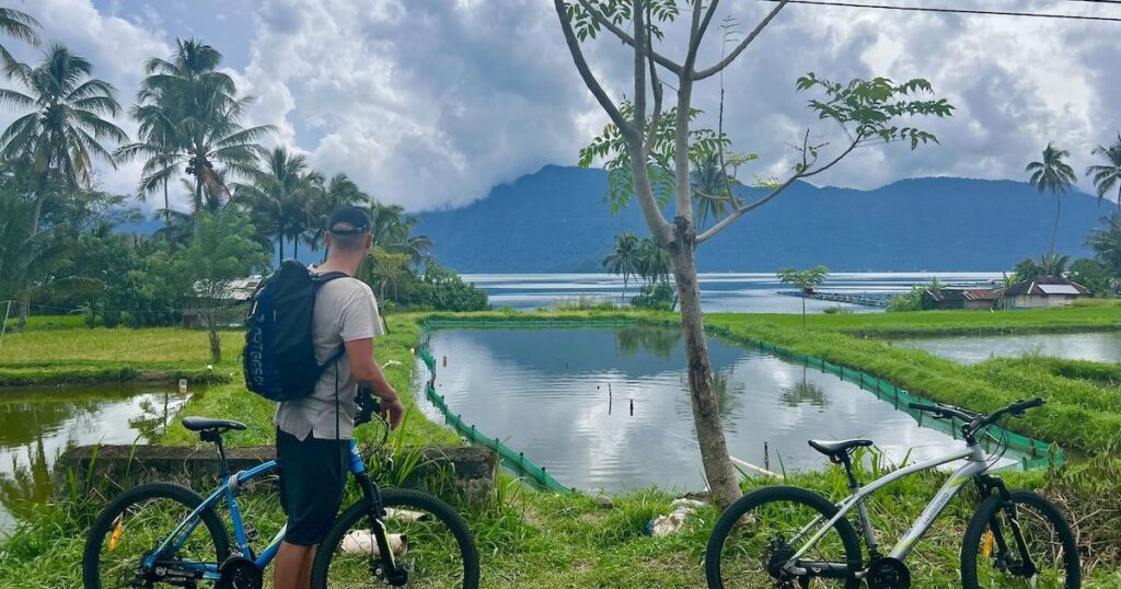 Man wearing a black cap, a grey t-shirt and long shorts holds a bicycle next to rice fields in Lake Maninjau.
