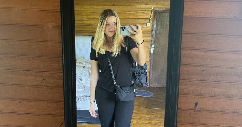 Women wearing a black t-shirt, black linen trousers and a black shoulder bag takes a selfie.