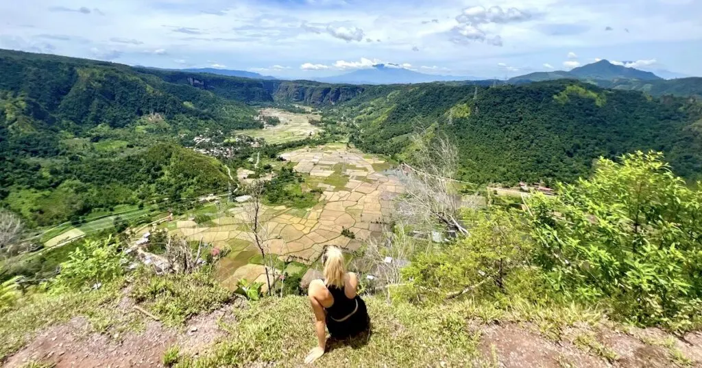 Female hiker sits on a dirt ledge above rice paddy fields and a volcano in Harau Valley in west Sumatra.