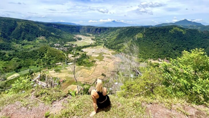 Female hiker sits on a dirt ledge above rice paddy fields and a volcano in Harau Valley in west Sumatra.