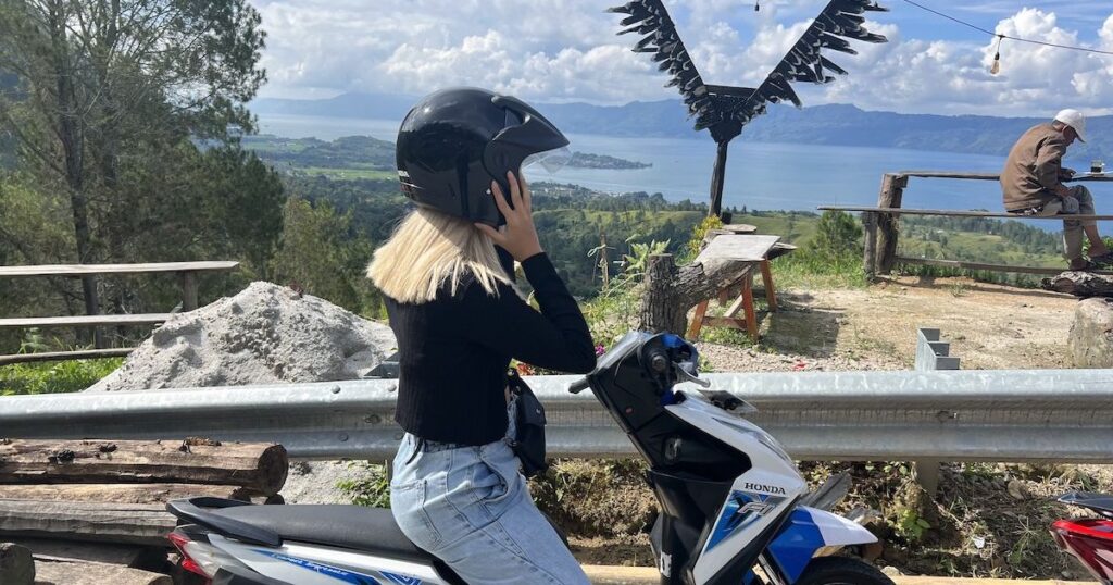 Women wearing blue denim jeans and a black cardigan clutches her scooter helmet on a road over Lake Toba.