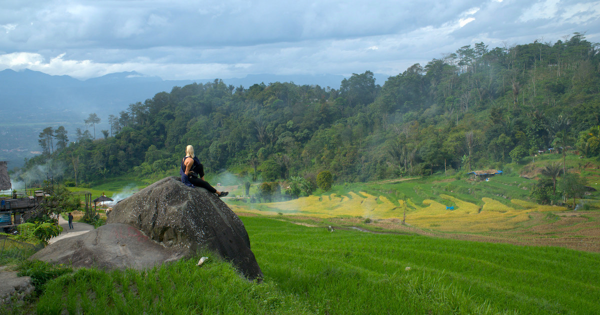 Woman wearing black linen trousers, a vest and a long-sleeved blue shirt sits on a rock overlooking rice terraces near Harau Valley.