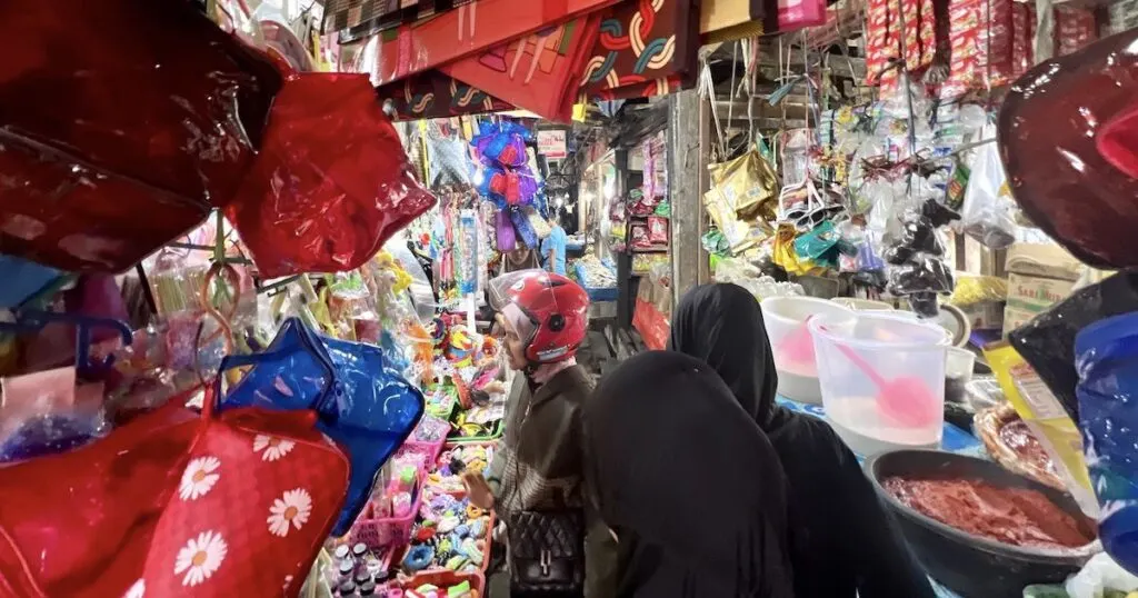 Women wearing hijabs walk through a narrow indoor market in Aur Kuning.