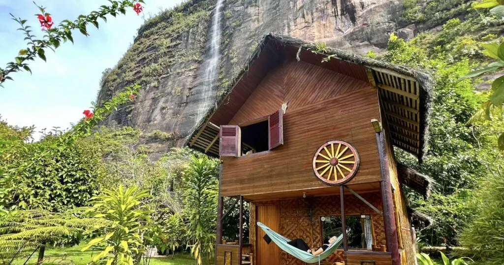 Woman lies in a hammock on the balcony of a hut at Abdi Homestay, with a waterfall in the background.