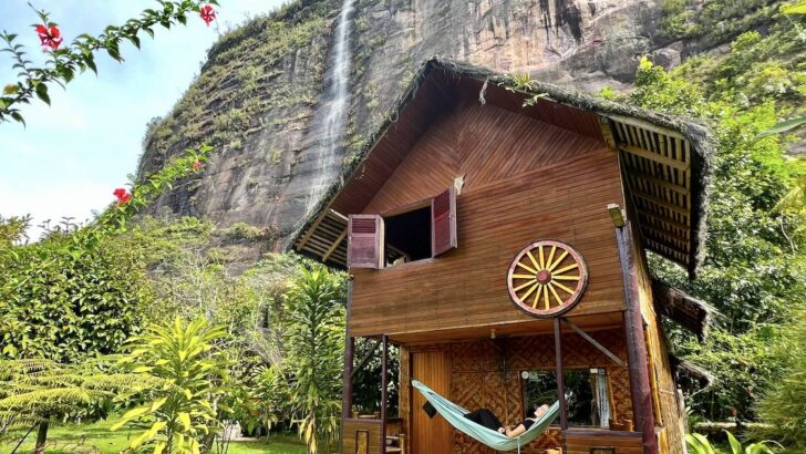 Woman lies in a hammock on the balcony of a hut at Abdi Homestay, with a waterfall in the background.