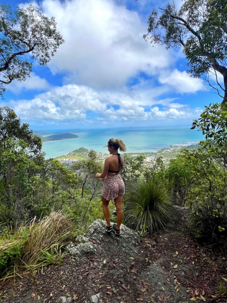 Woman stands at the Honeyeater Trail Lookout with Airlie Beach and the Whitsunday Islands in the background.