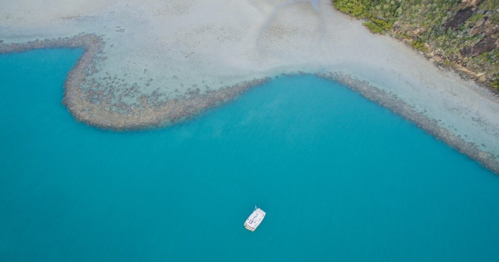 Aerial view of a lone white sailing boat floats just off shore in the Whitsundays.