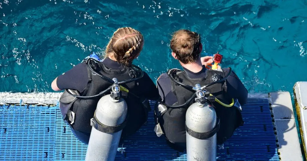 Two scuba divers with tanks sit on a platform above the Cairns Barrier Reef.