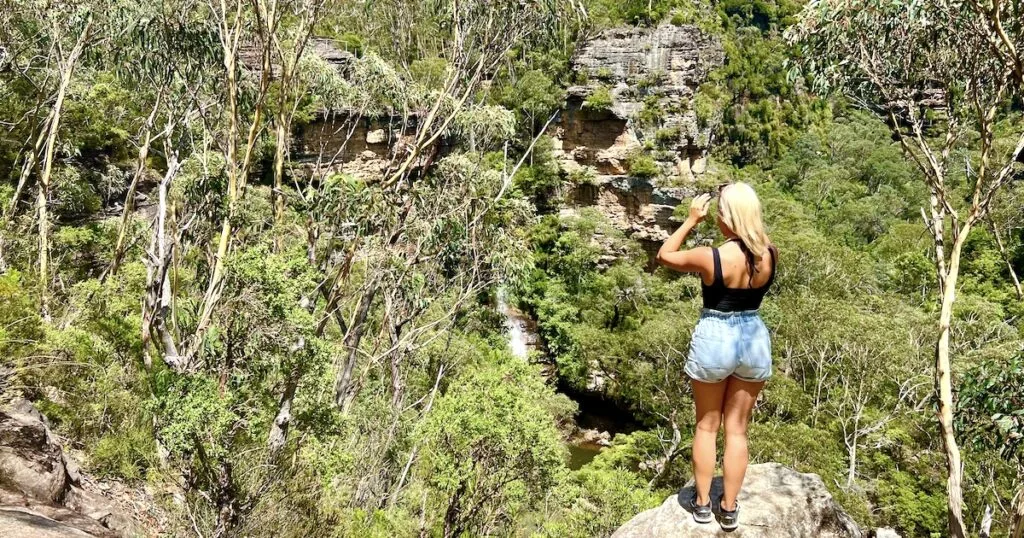 Woman stands on a rock overlooking dense bush surrounding the tall Minnehaha Falls in the Blue Mountains.