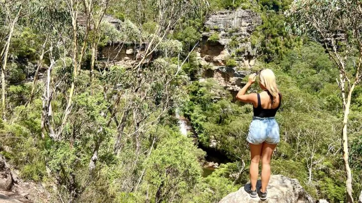 Woman stands on a rock overlooking dense bush surrounding the tall Minnehaha Falls in the Blue Mountains.