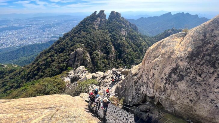 Hikers ascend steep stairs alongside boulders on Bukhansan Mountain, with views of a rocky, green peak in the background.
