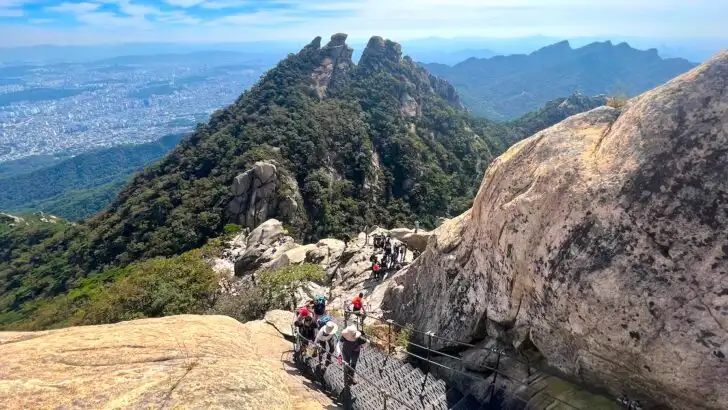 Hikers ascend steep stairs alongside boulders on Bukhansan Mountain, with views of a rocky, green peak in the background.