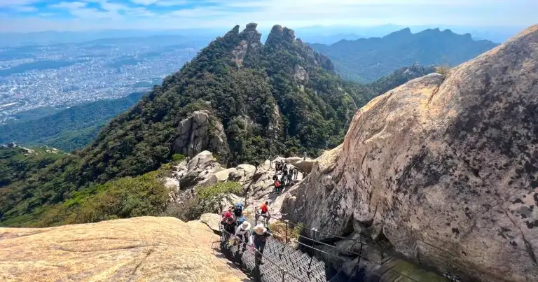 Hikers ascend steep stairs alongside boulders on Bukhansan Mountain, with views of a rocky, green peak in the background.