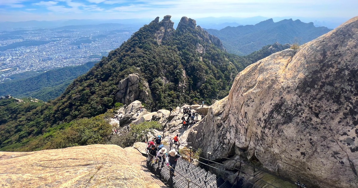Hikers ascend steep stairs alongside boulders on Bukhansan Mountain, with views of a rocky, green peak in the background.