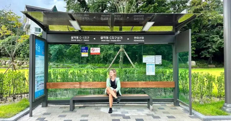 Woman wearing a green raincoat sits on a bench at the Seorak-dong C Shopping District bus stop near Seoraskan National Park.