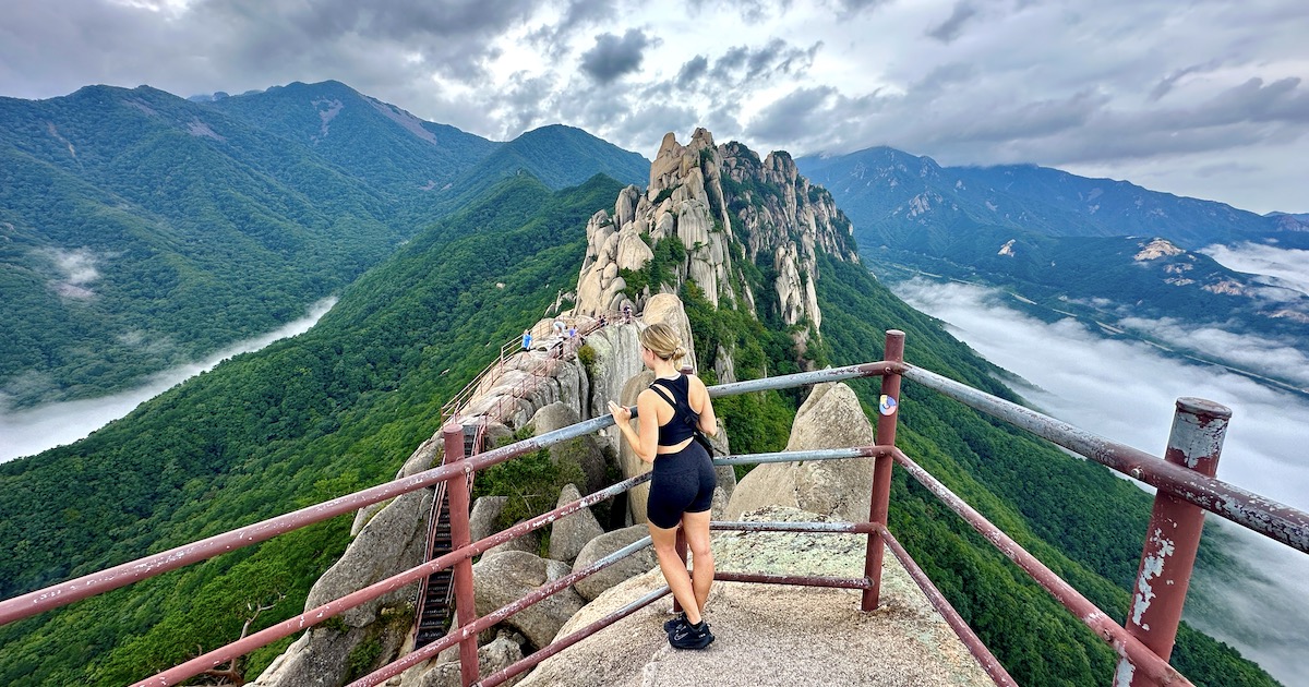 Woman wearing hiking gear gazes over Ulsanbawi's granite boulders and a green valley.
