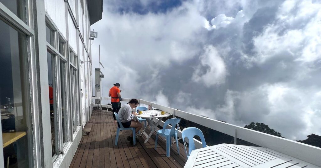 Hikers sip on coffee on plastic chairs at the Laban Rata Rest House balcony, overlooking white, fluffy clouds.
