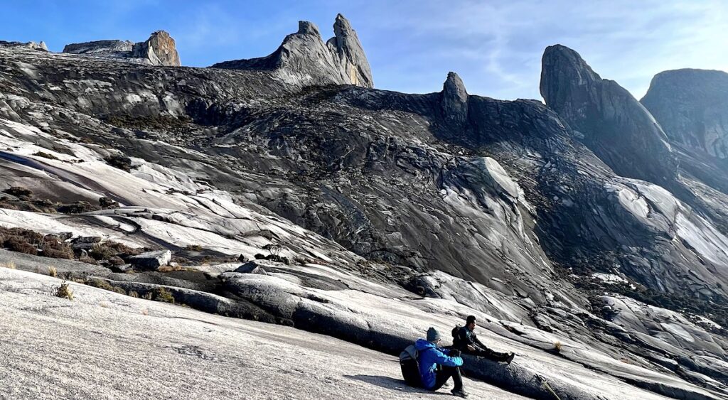 Two hikers rest on a sloping rocky plain at Mount Kinabalu, with dramatic rocky peaks in the distance.