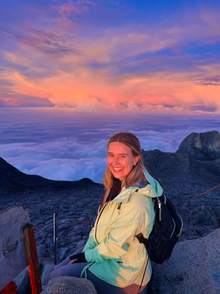 A woman wearing a green rainjacket smiles at the top of Mount Kinabalu, with the sunrise turning the clouds pink and purple.