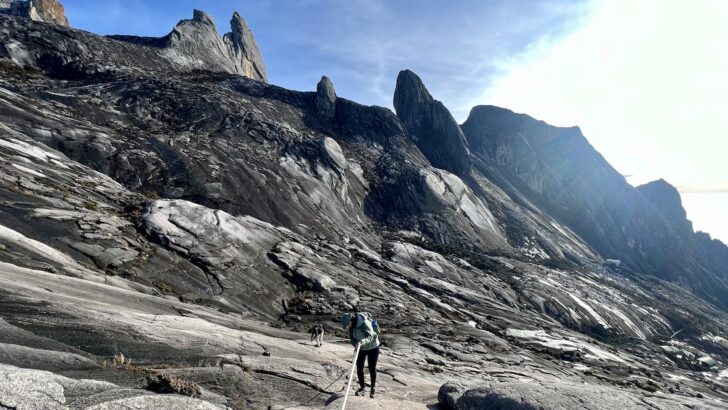 A female hiker uses ropes to descend down a rocky surface near the summit of Mount Kinabalu, with large, sharp boulders in the background.