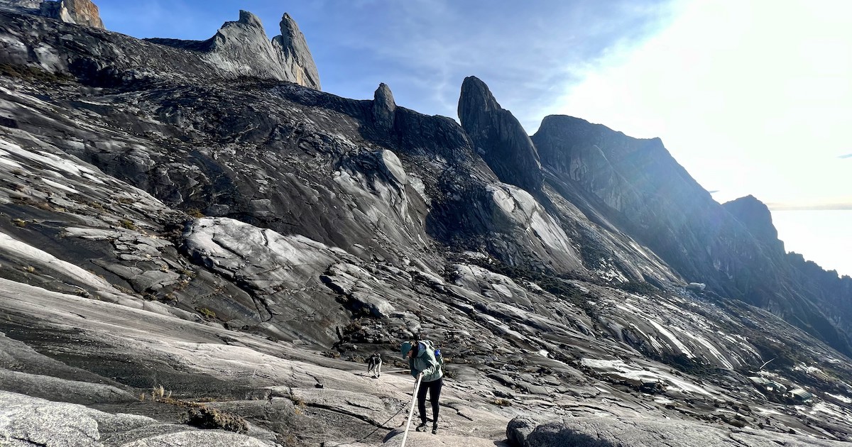 A female hiker uses ropes to descend down a rocky surface near the summit of Mount Kinabalu, with large, sharp boulders in the background.