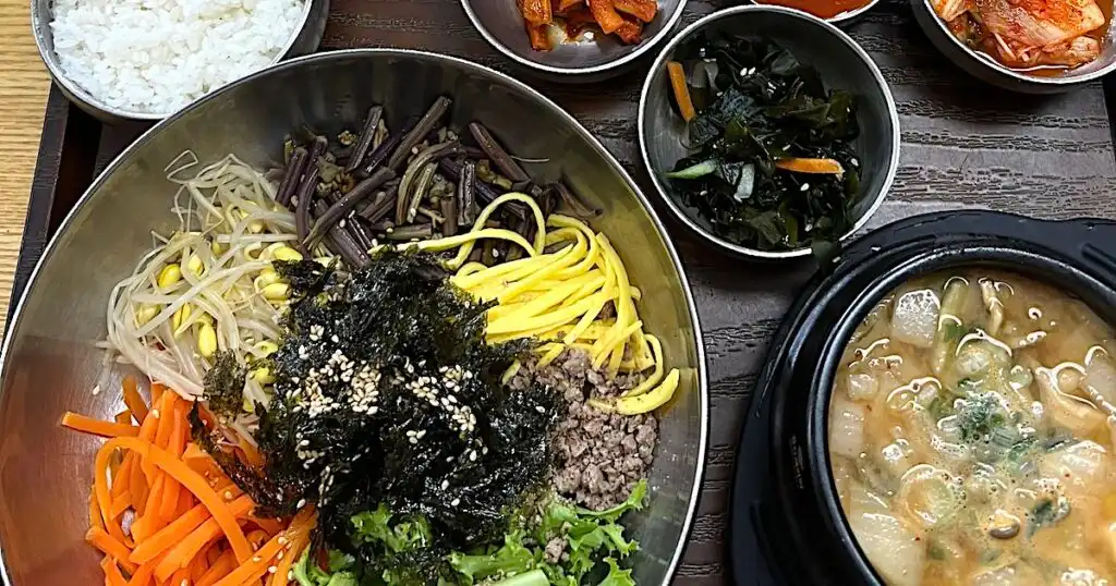 A round bowl containing Korean bibimbap, accompanied by a soup, kimchi, white rice and other sides.