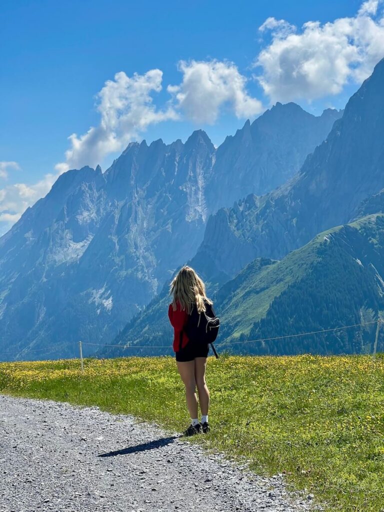 Woman standing beside a mountain trail looking towards dramatic jagged Alpine peaks.