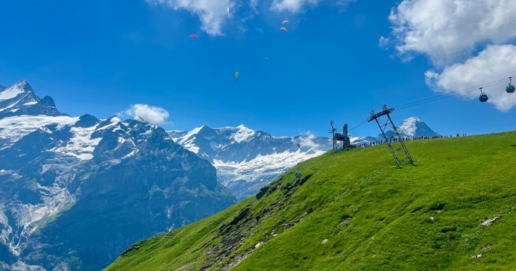 Snow-capped Alpine mountains overlooking a green hillside with cable cars and paragliders soaring overhead.